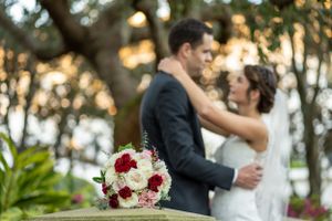 Romantic wedding couple embracing outdoors with red and white bouquet at sunset.