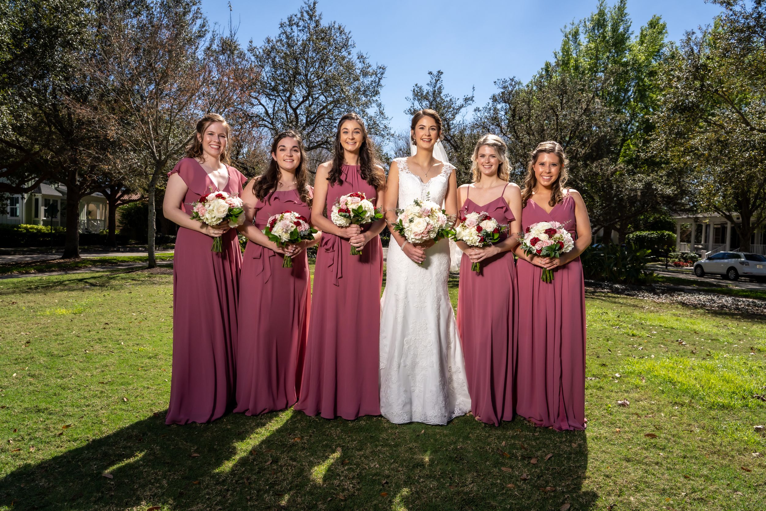 Bridesmaids in burgundy dresses stand with bride on grassy lawn for wedding photo.