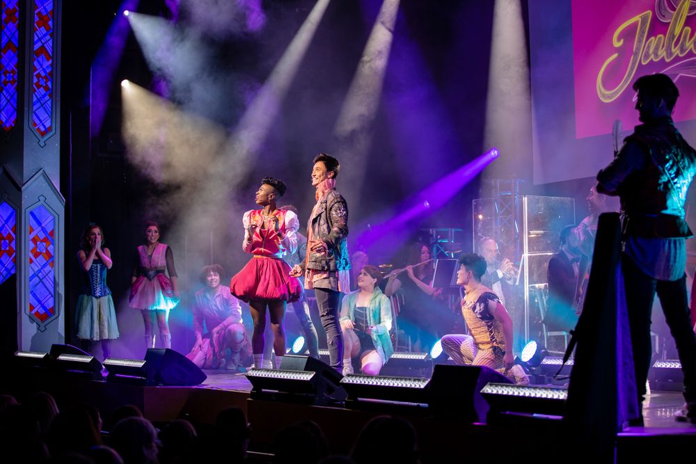Performers in retro costumes dance on stage with purple and pink theatrical lighting effects.