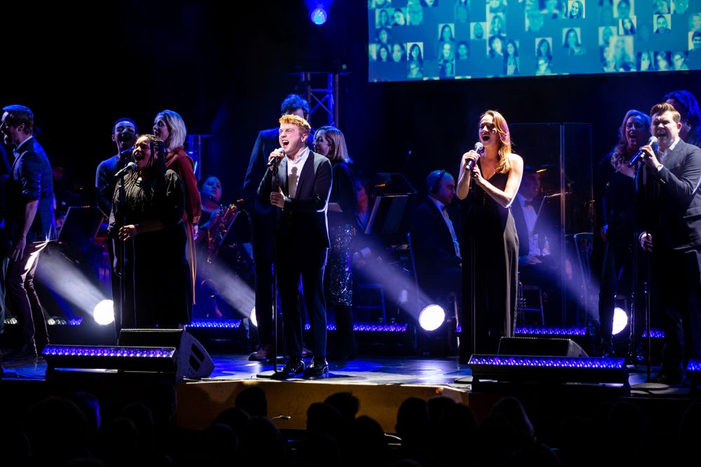Performers singing on stage with dramatic blue spotlights and video screen backdrop.