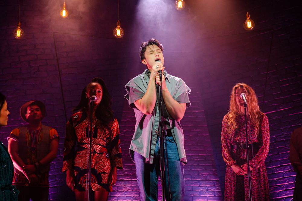 Performance ensemble on stage with purple theatrical lighting against a brick wall backdrop.