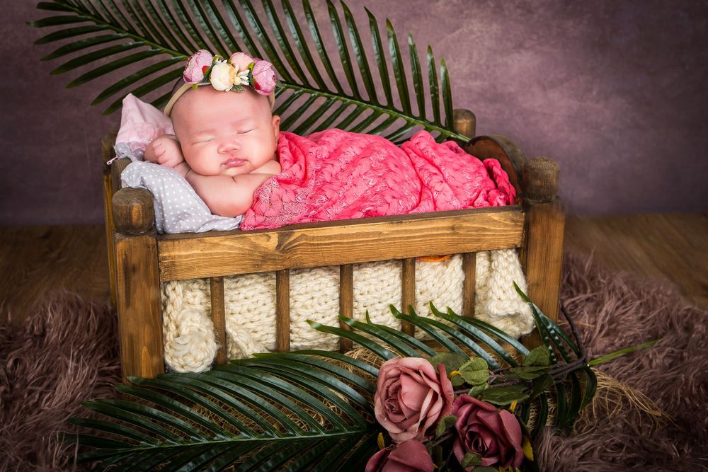 Newborn baby girl sleeping in a rustic wooden bed, wrapped in a pink blanket with floral decor. Cozy newborn photography session