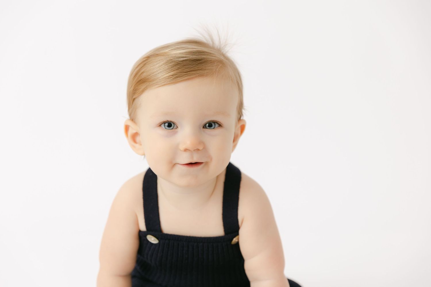 Seven month old baby with blonde hair and blue eyes smiles at the camera. Baby is wearing a navy romper with thin straps