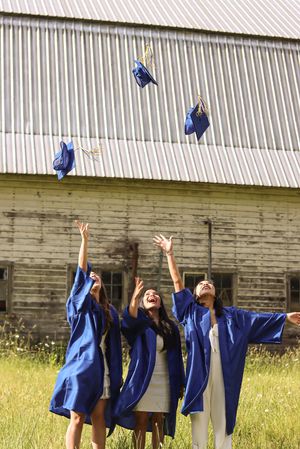 Cora Emilie Media Graduates throwing blue caps in the air in front of rustic barn building.