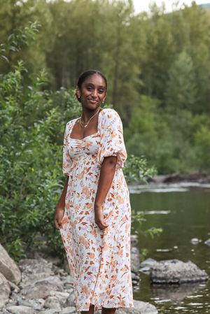 Cora Emilie Media A summer portrait in a white floral dress standing on rocks by water.