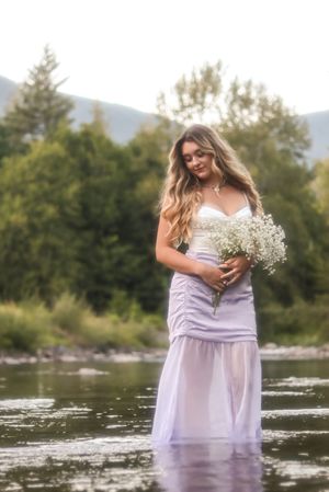 Cora Emilie Media A dreamy portrait in a lavender dress holding baby's breath flowers by a mountain stream.
