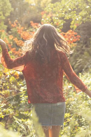 Cora Emilie Media Person in red top and denim skirt dances in sunlit autumn garden with golden foliage.