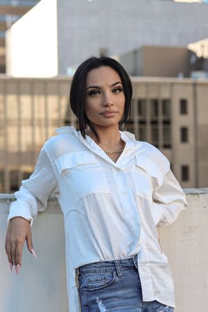 Cora Emilie Media Woman in white button-down shirt and jeans poses on urban rooftop.