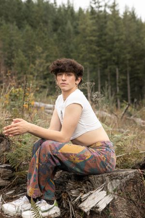 Cora Emilie Media A person in white crop top and patterned pants sits on a log in a forest setting.