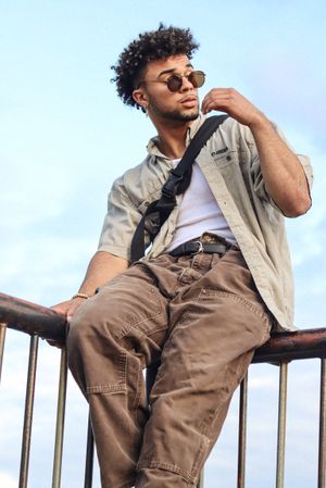 Cora Emilie Media Stylish man in casual wear and sunglasses posing on railing against blue sky.