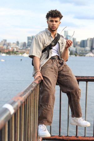 Cora Emilie Media Urban fashion shot of man in beige shirt and cargo pants leaning on waterfront railing.