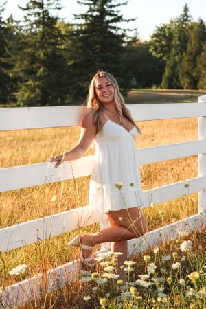 Cora Emilie Media A casual portrait by a white fence surrounded by wildflowers at golden hour.
