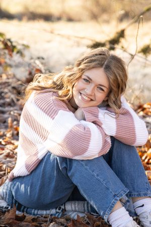 Cora Emilie Media Person in pink sweater and jeans sits relaxed on fallen leaves during autumn photoshoot.