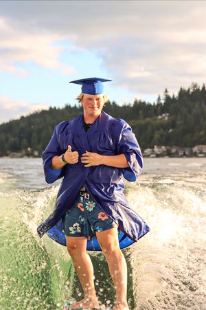 Cora Emilie Media Graduate in blue cap and gown poses in water during celebration photoshoot.