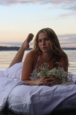 Cora Emilie Media Figure reclines on white bedding by water holding baby's breath flowers at dusk.