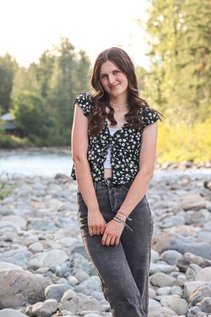 Cora Emilie Media A portrait shot beside a rocky river showing a floral top and dark jeans in a natural outdoor setting.