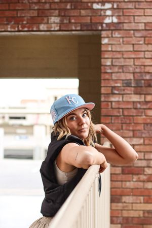 Cora Emilie Media Woman in baseball cap and casual wear leans against railing by brick wall.