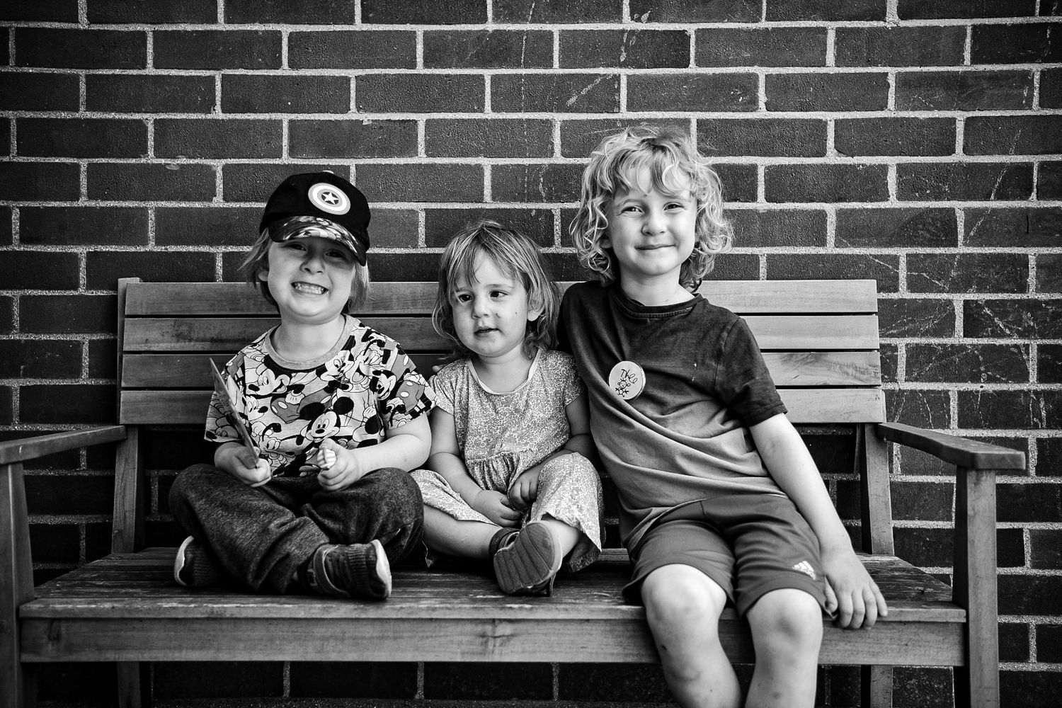 two boys and their little sister sit together in a row on a bench smiling at the camera