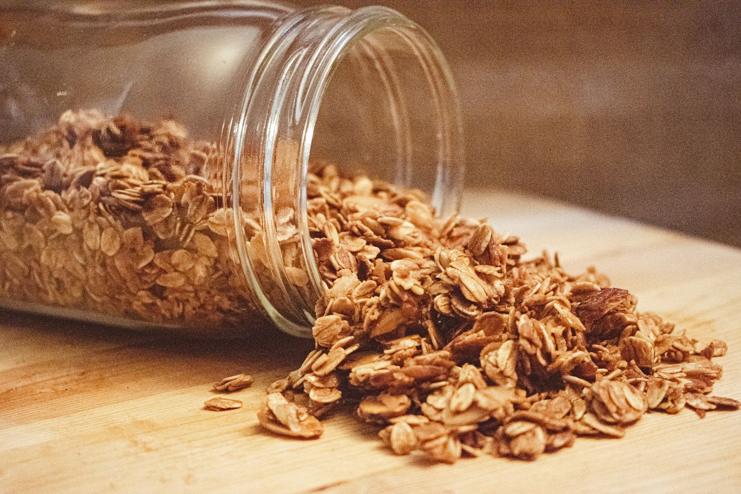 Homemade granola spilling from a glass mason jar onto a wooden surface in warm lighting.