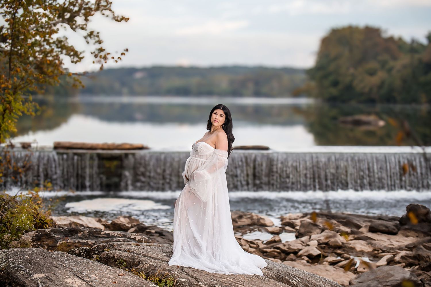 Person in flowing white dress poses on rocky waterfall ledge during maternity photoshoot.