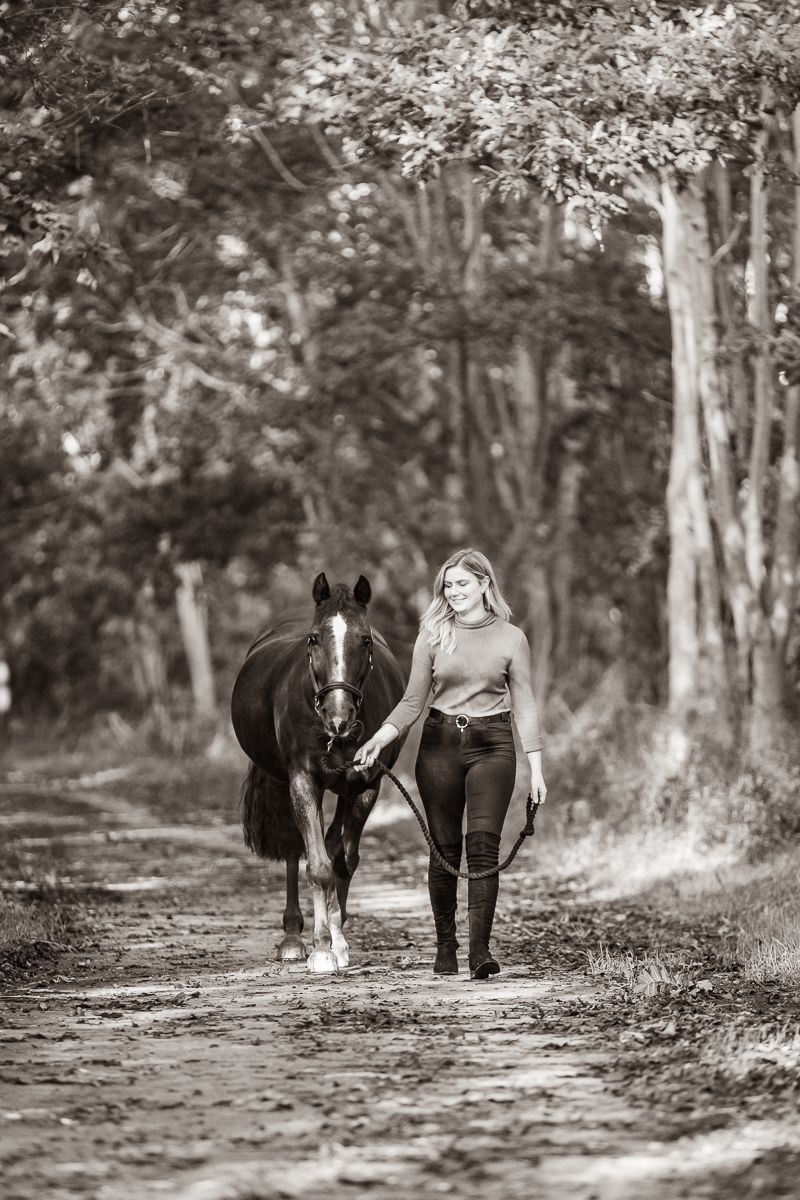 A black and white photo shows a person walking with their horse down a tree-lined path.