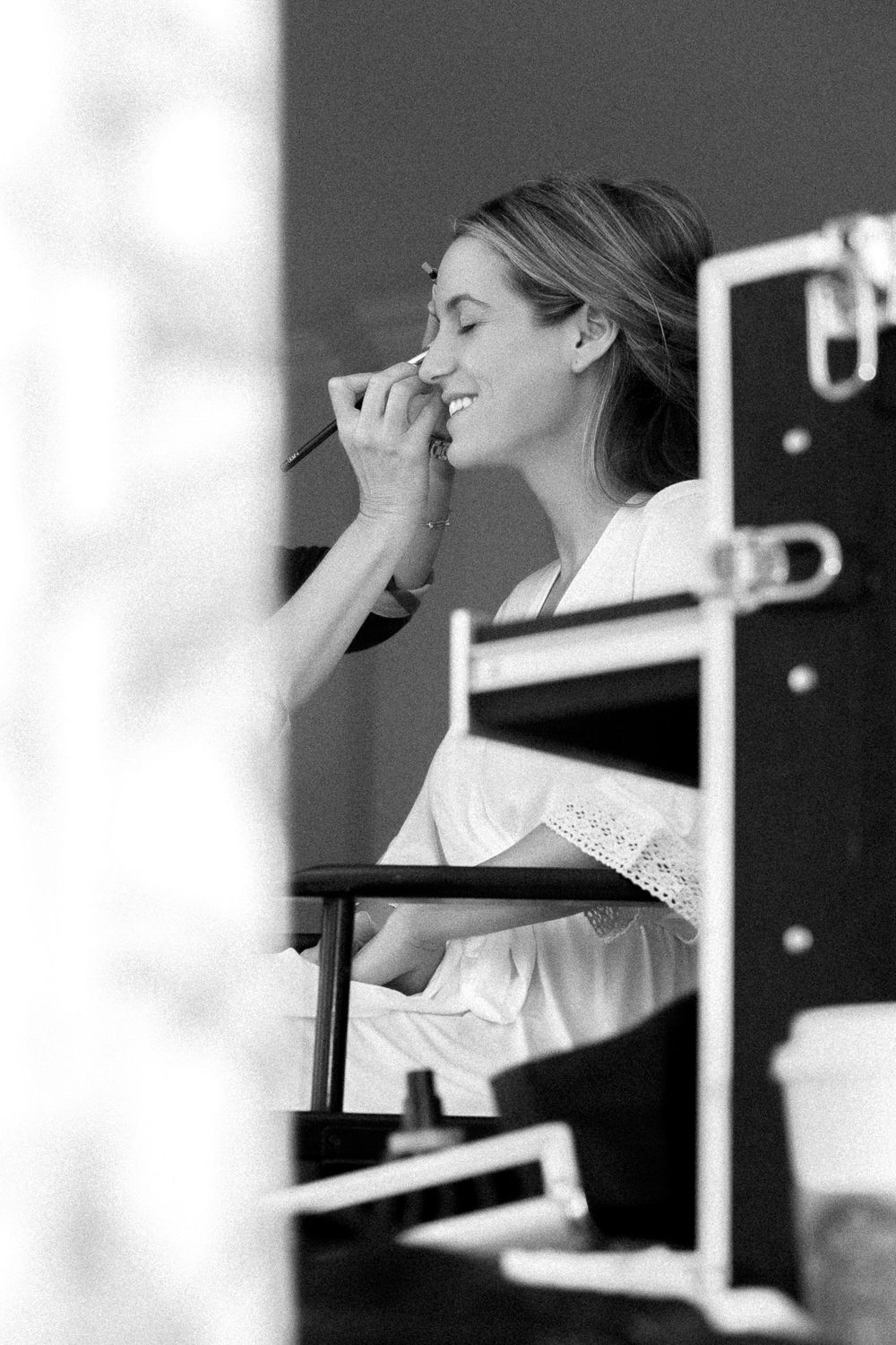 Black and white profile photo of someone in a director's chair getting makeup applied while wearing a white robe.