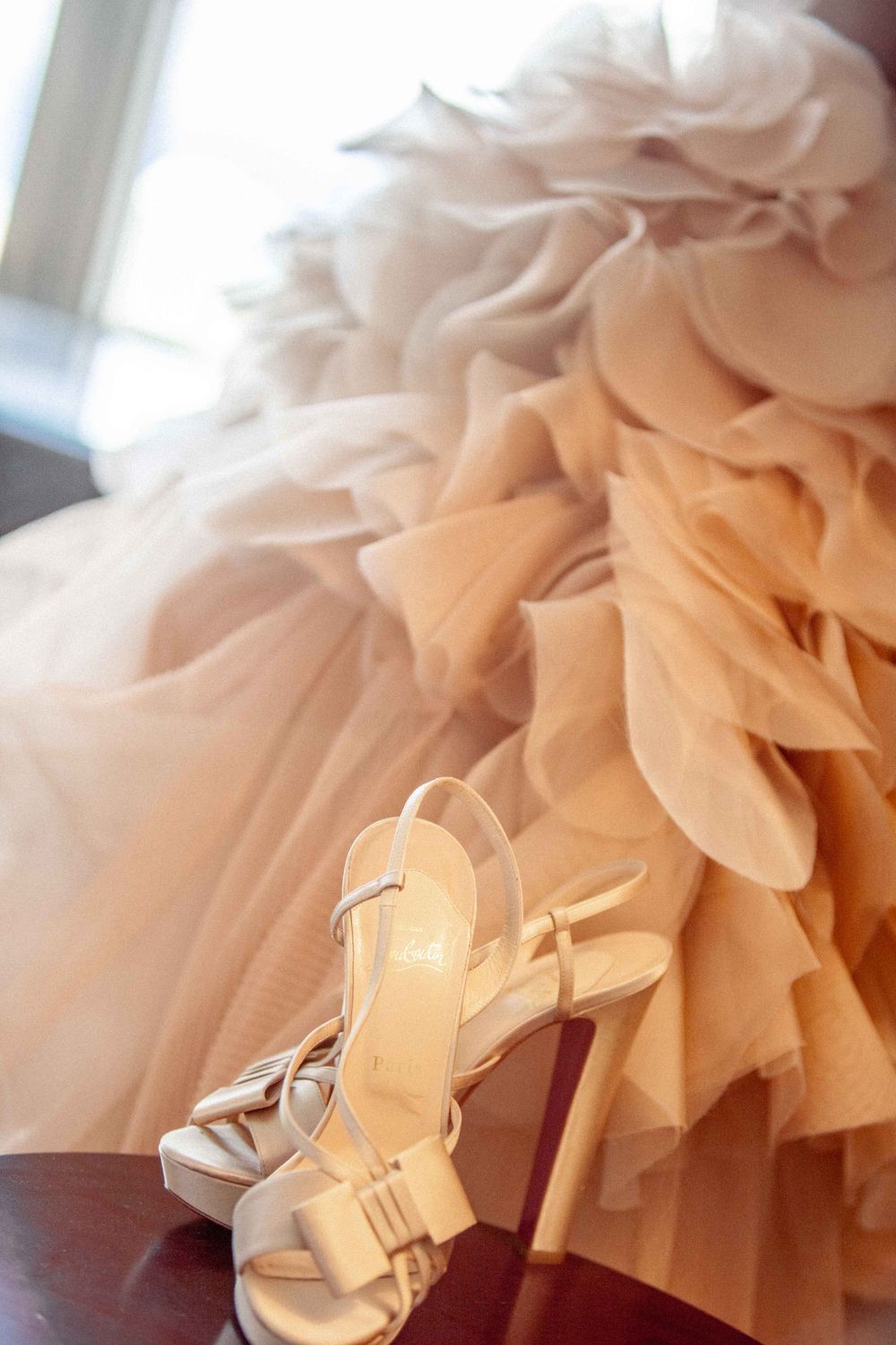 Elegant peach tulle wedding dress fabric and white high heel bridal sandals with bow detail rest on wooden surface.