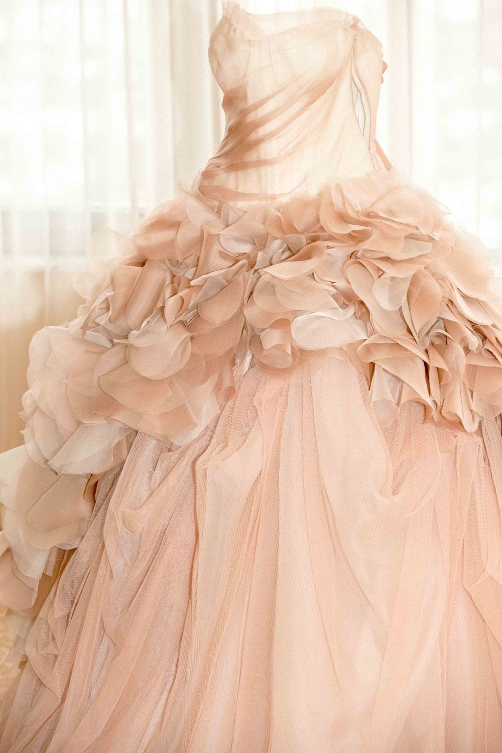 A blush pink wedding gown with ruffled tulle details and a strapless bodice hangs elegantly against sheer curtains.