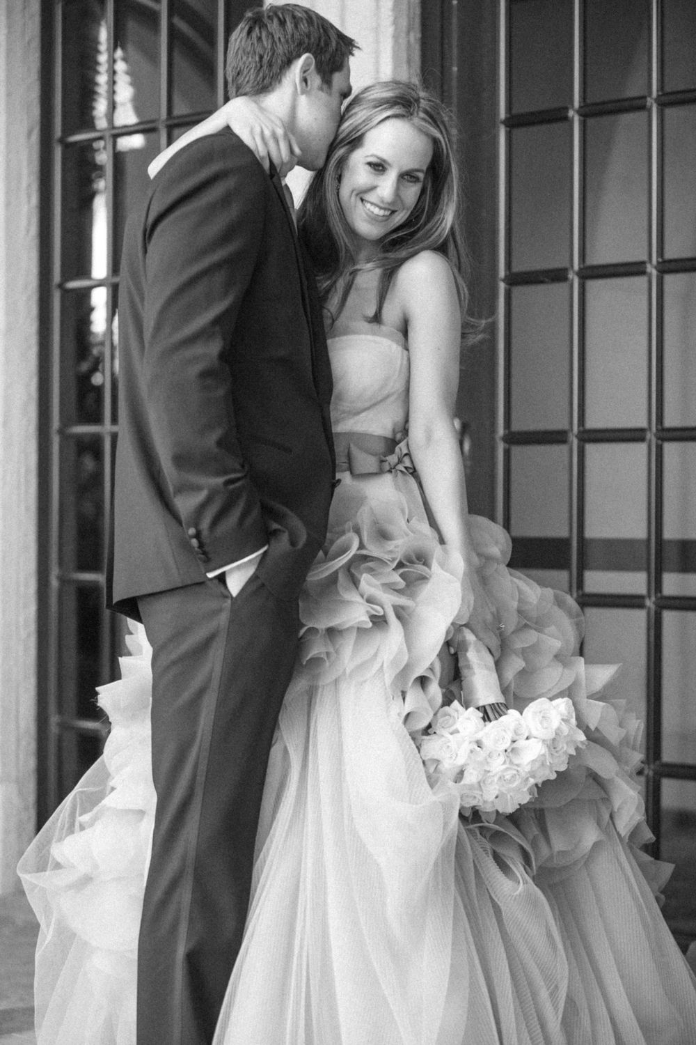 Bride in flowing white dress and groom in dark suit share intimate wedding moment in front of industrial windows.