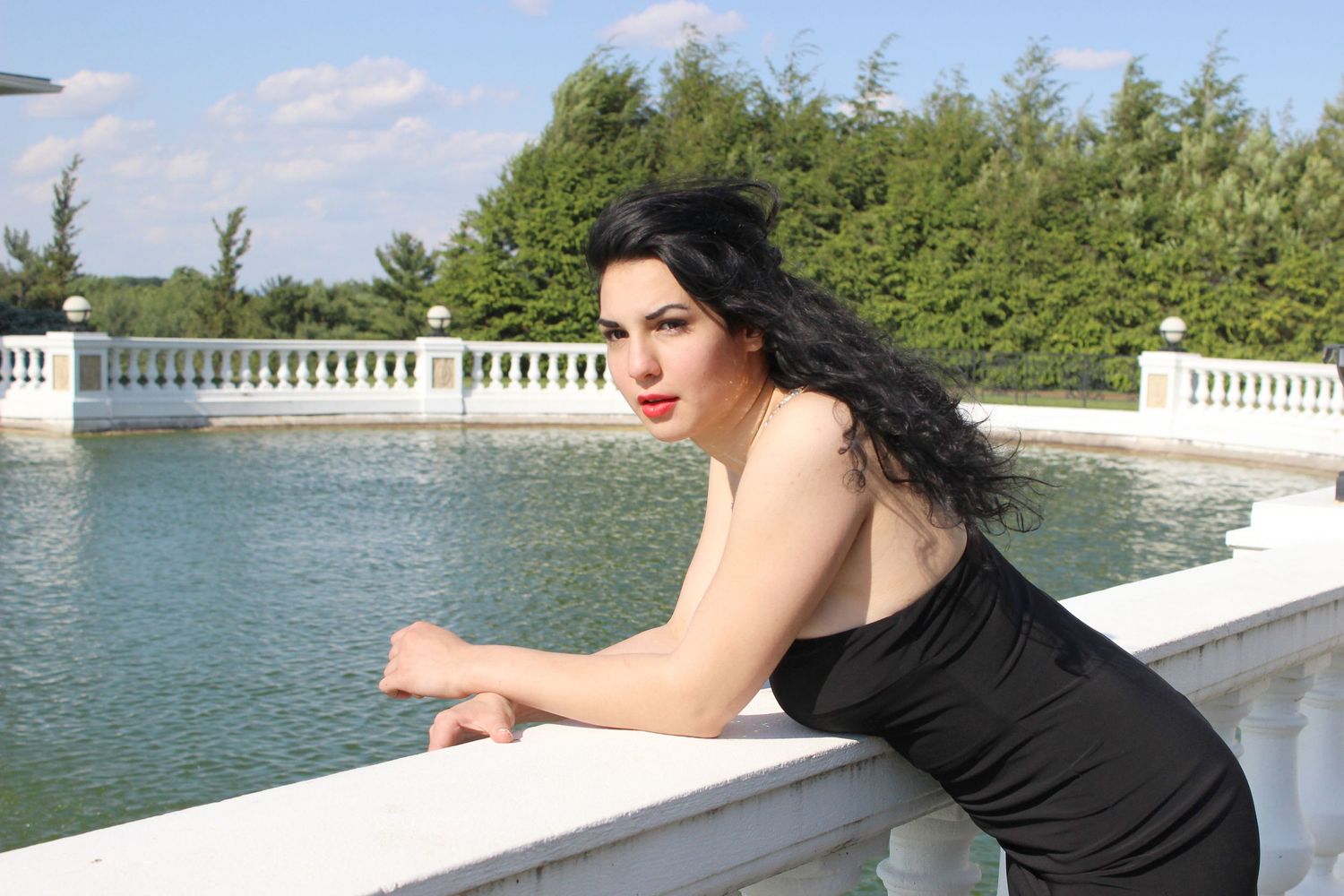 Woman in black dress leaning on white balustrade near ornamental pond.