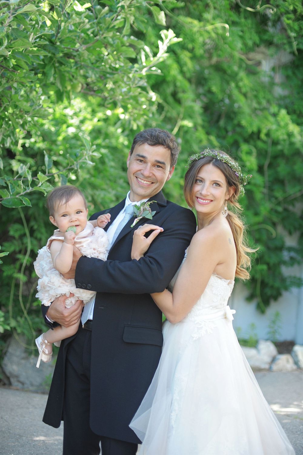 A wedding couple in formal attire poses outdoors with a baby against a green garden backdrop at God's Mountain, Penticton B.C.