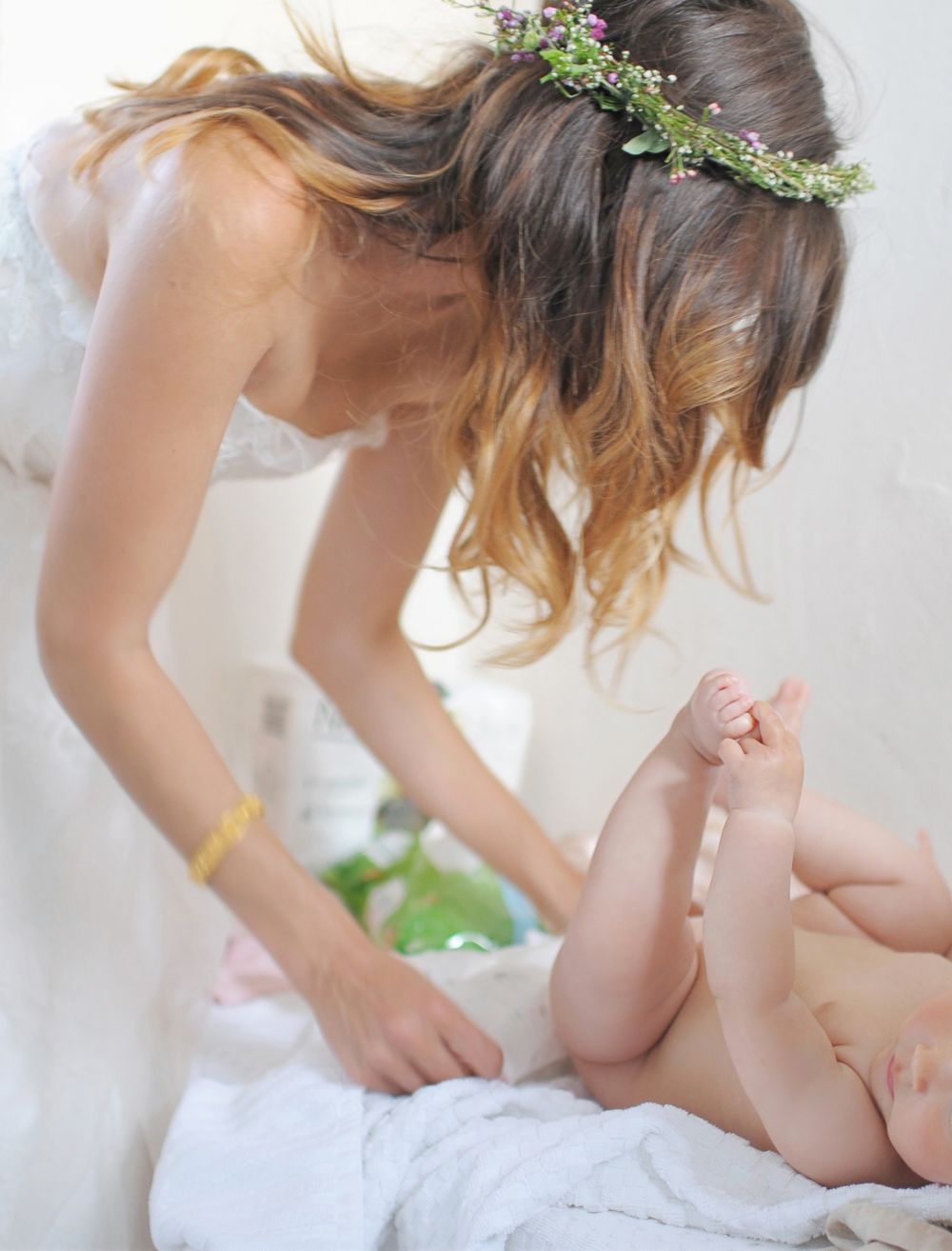 A bride in a floral crown leans over a baby lying on white bedding at God's Mountain, Penticton B.C.