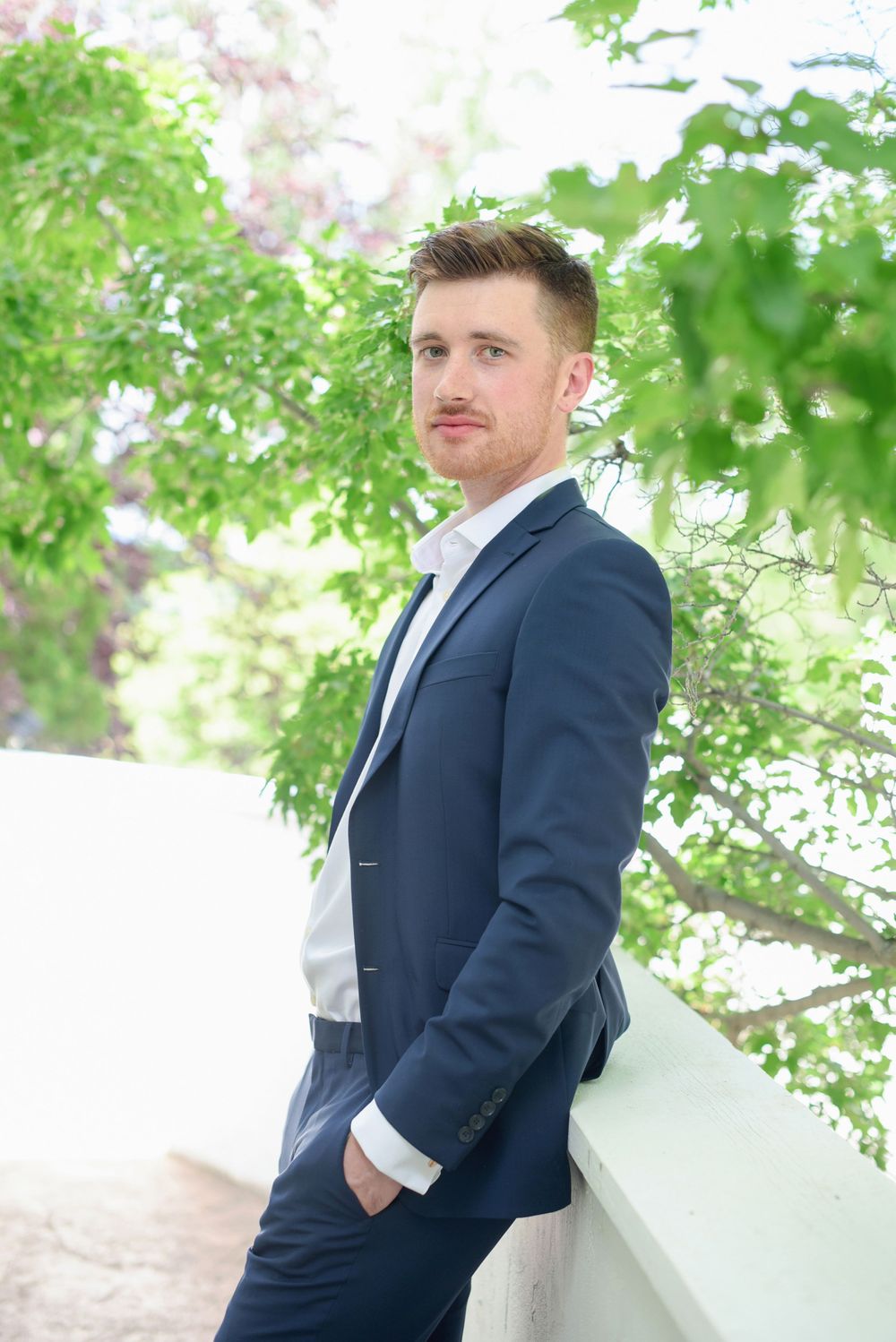 Groom in navy suit poses against spring blossoms and greenery at God's Mountain, Penticton B.C.