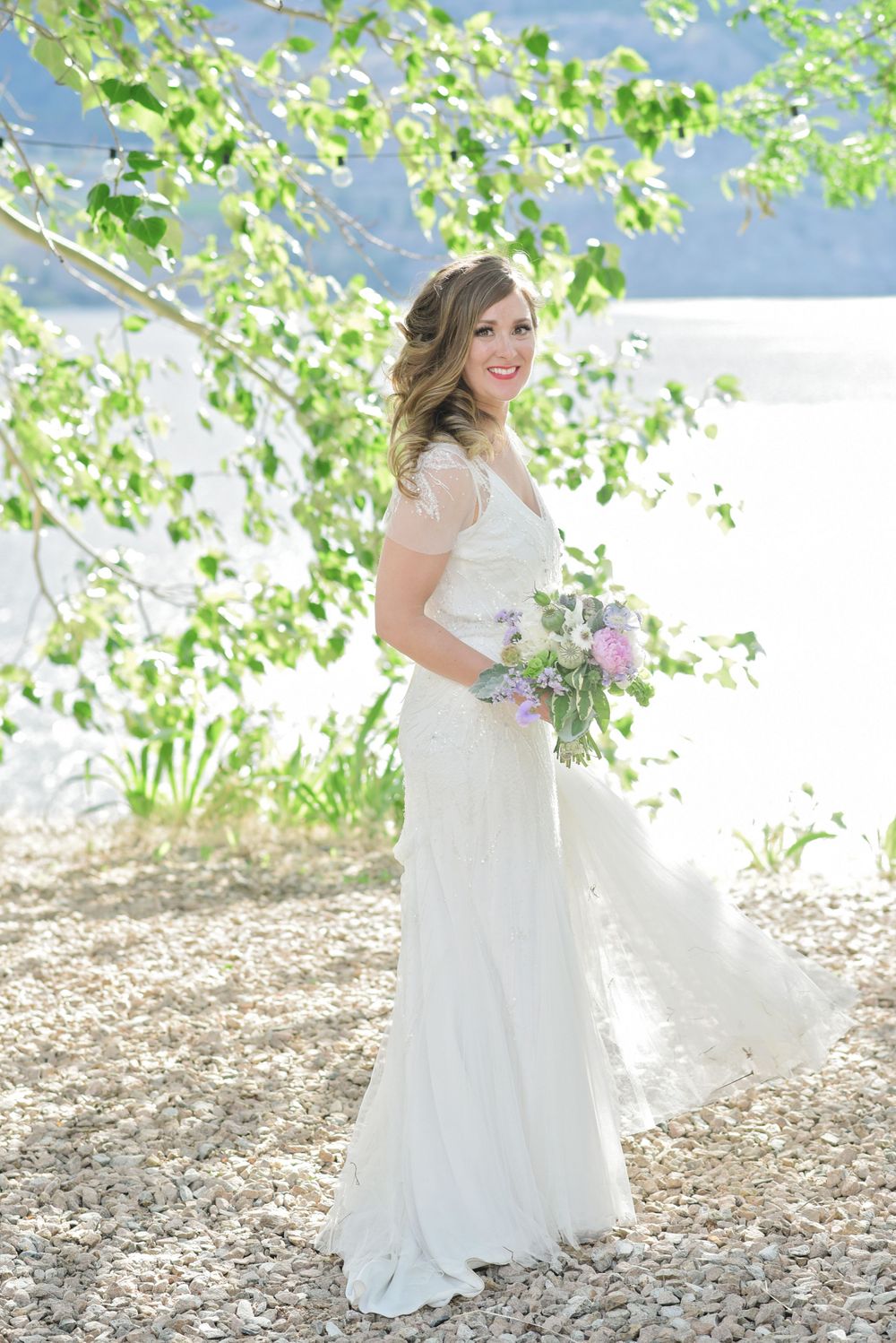 Bride in flowing white dress poses with bouquet against lakeside greenery at God's Mountain, Penticton B.C.