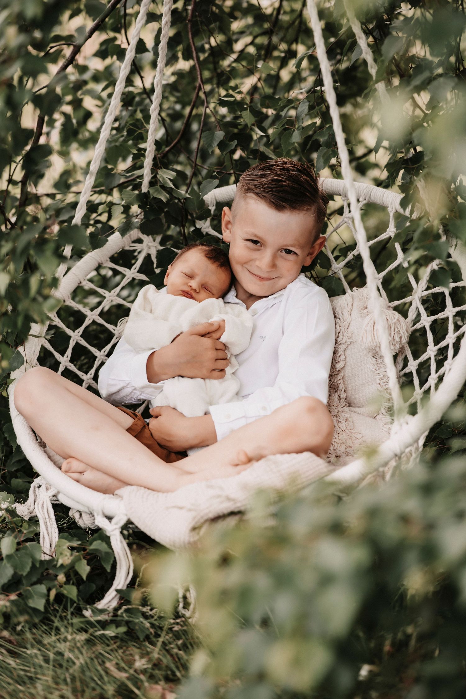 Two siblings share a tender moment relaxing together in a macrame hanging chair surrounded by greenery.