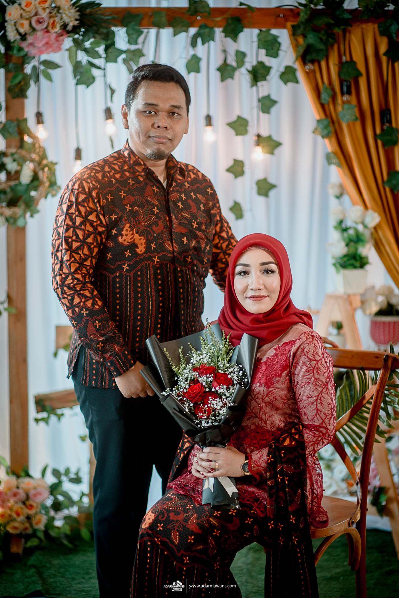 Couple in matching batik attire poses with bouquet against floral wedding backdrop.