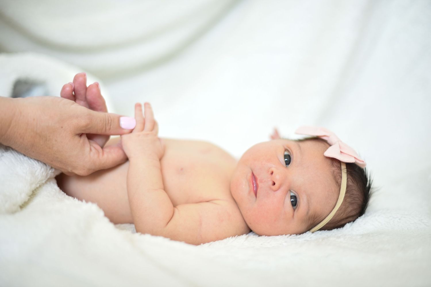 Alert newborn baby wearing white headband lying on soft white fabric.