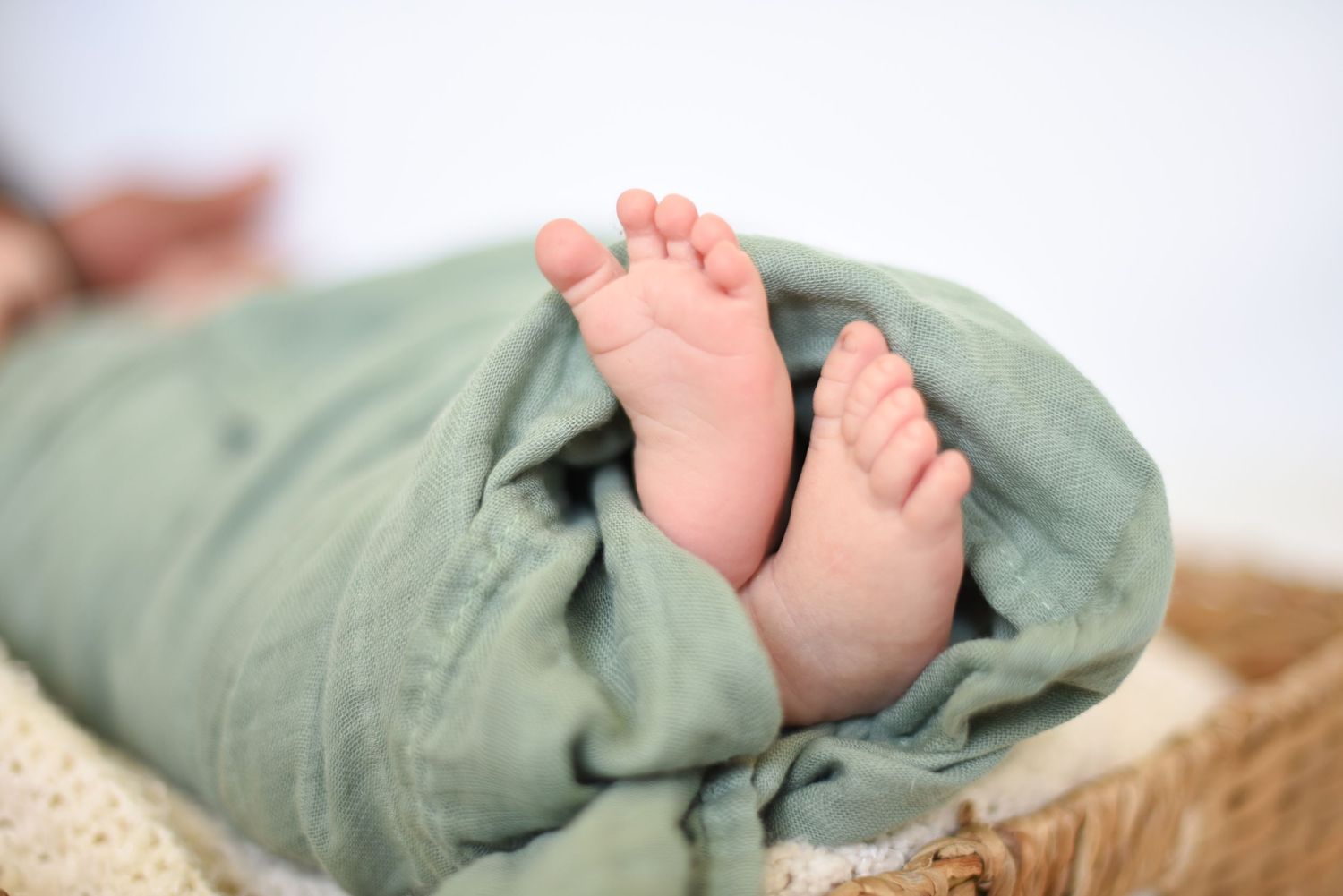 Tiny baby feet wrapped in soft sage green blanket nestled in woven basket.