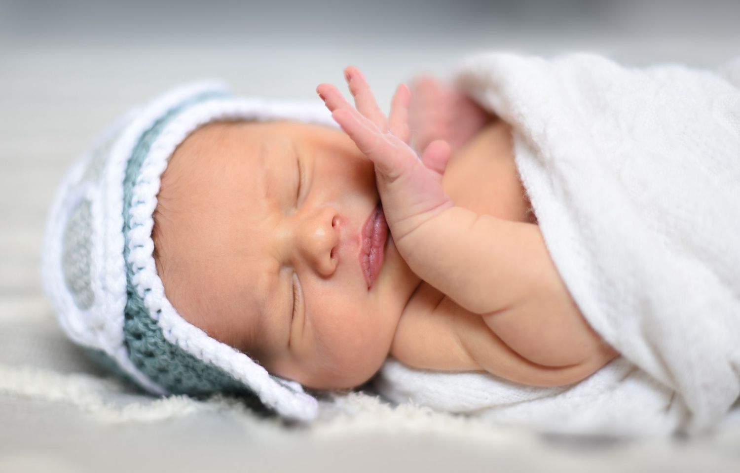 Peaceful sleeping baby wearing soft blue knit hat wrapped in white blanket.