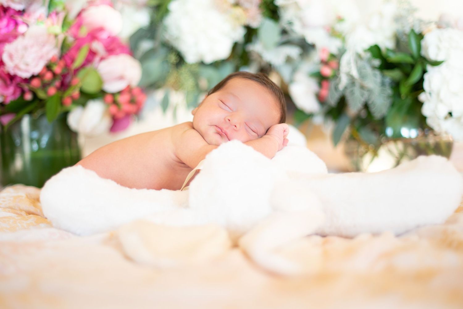 Peaceful newborn sleeping on cream blanket surrounded by floral arrangements.
