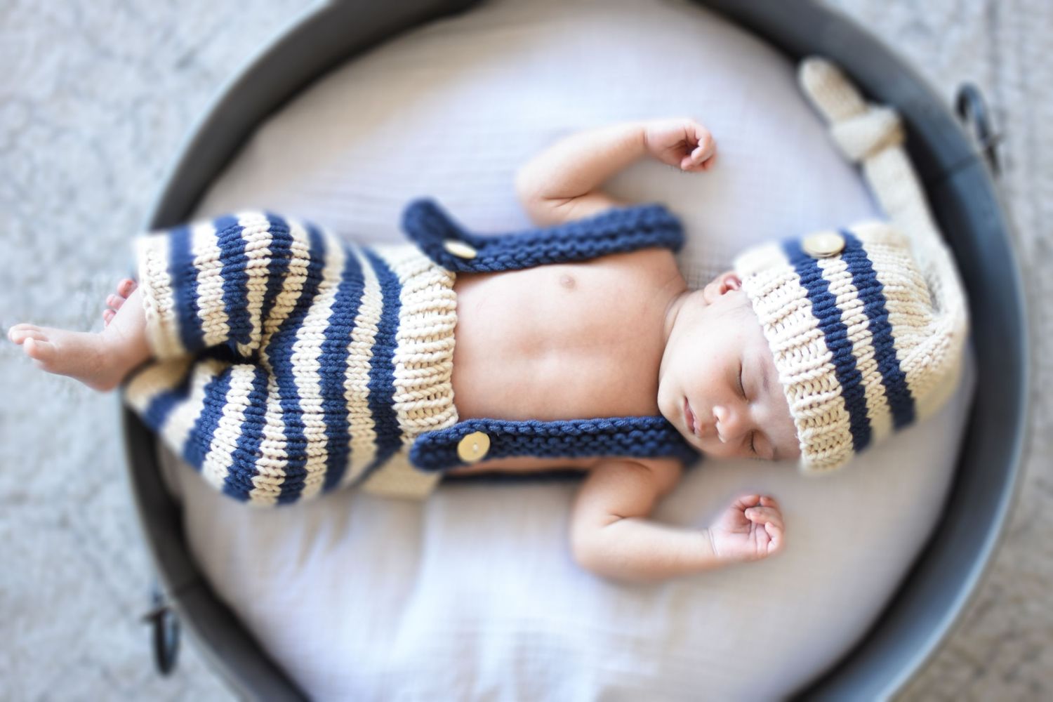 Newborn baby sleeping in gray basket wearing striped knit outfit.