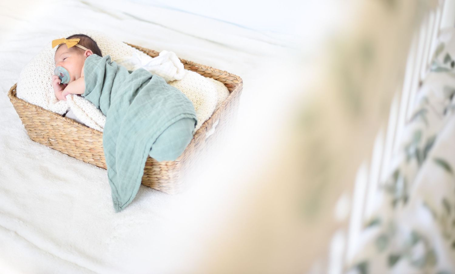 Tiny baby wearing mint green swaddle sleeping in woven basket on white surface.