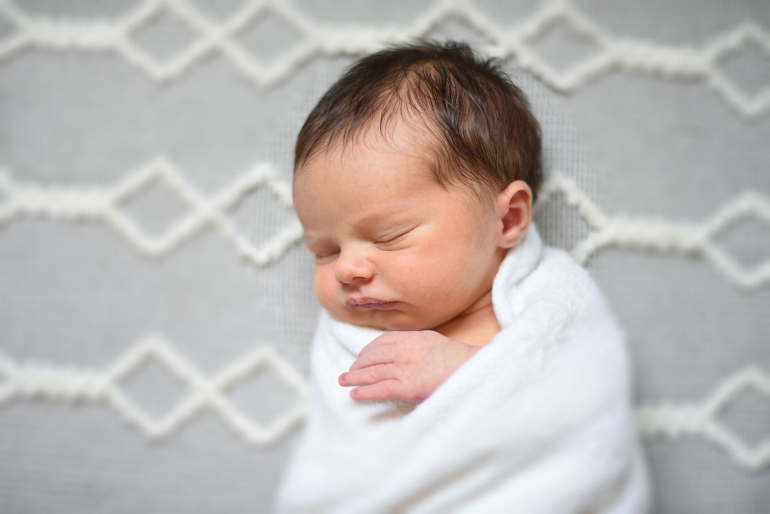 Peaceful newborn baby swaddled in white blanket sleeping against gray patterned background.