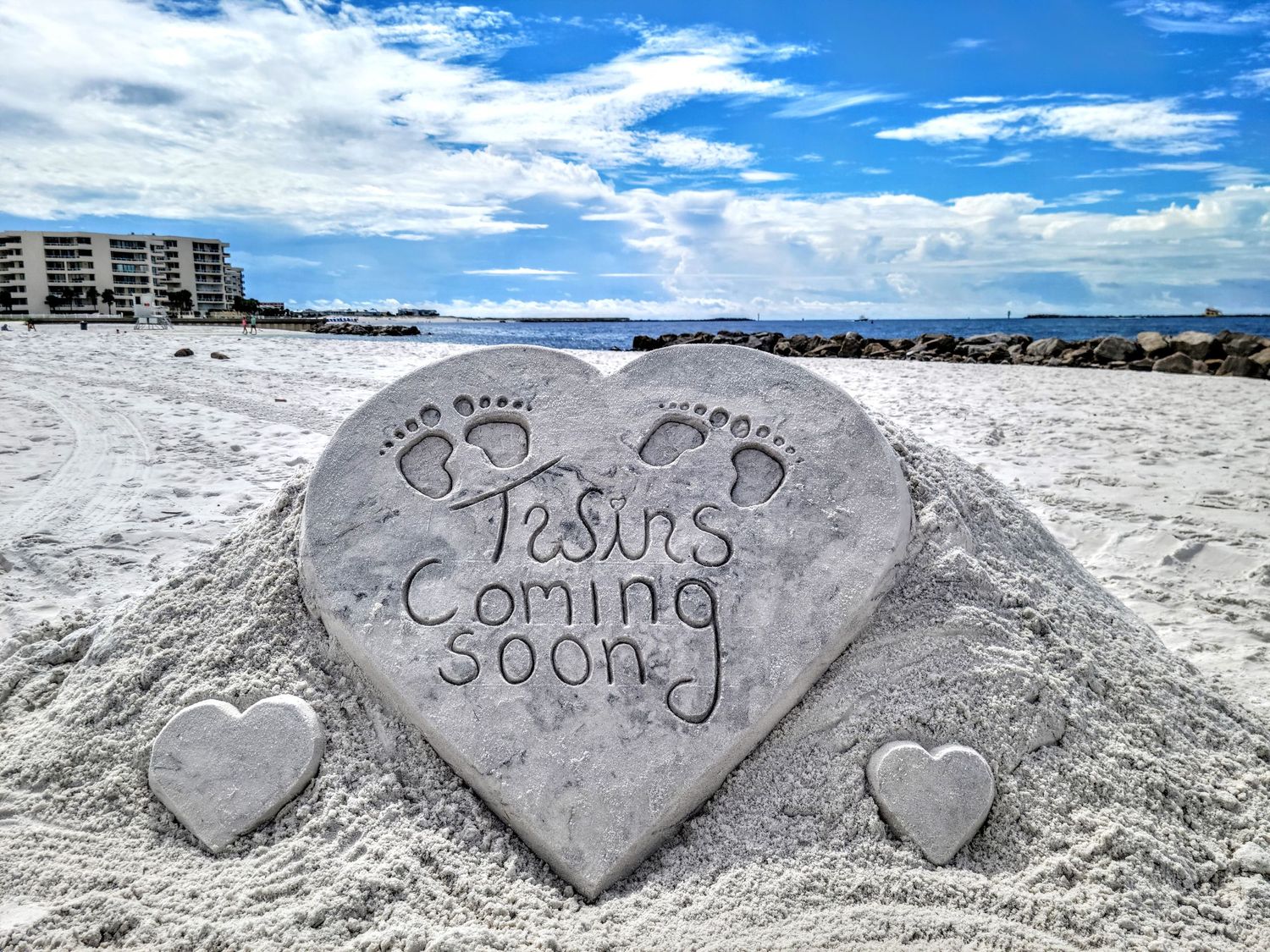 A heart-shaped sand sculpture on a beach with 'Twins coming soon' message and baby footprints carved into it.