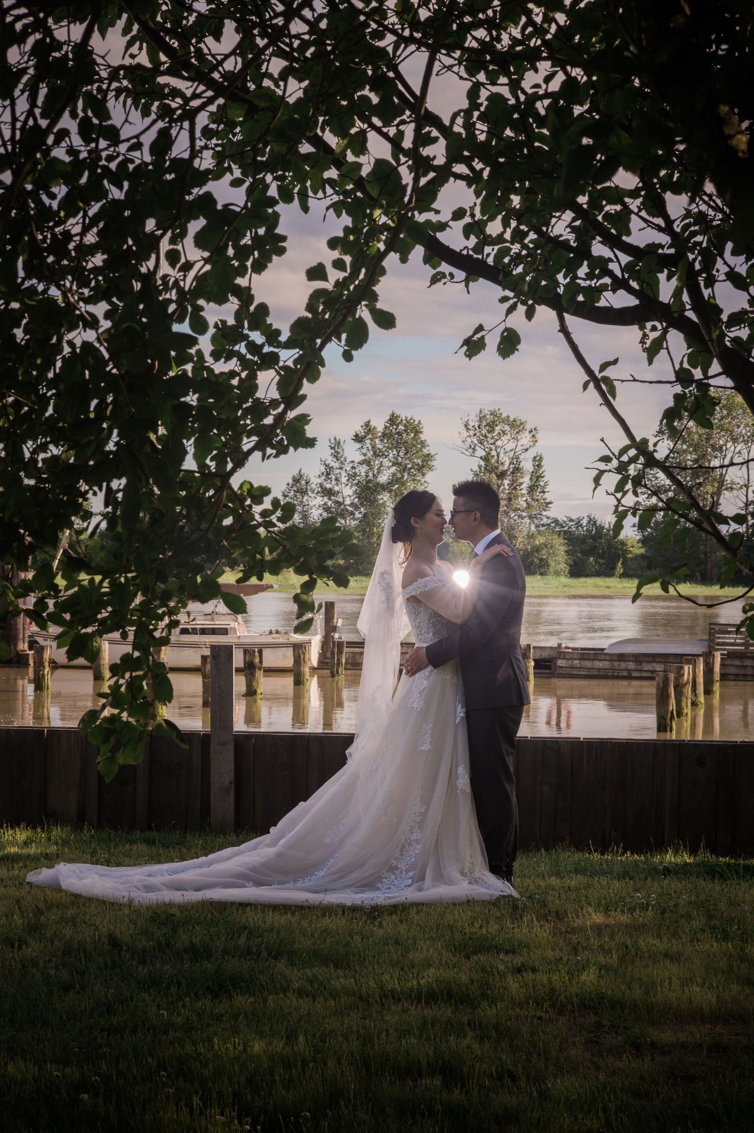 A romantic wedding couple silhouetted against sunlight by a waterfront dock with overhanging tree branches.