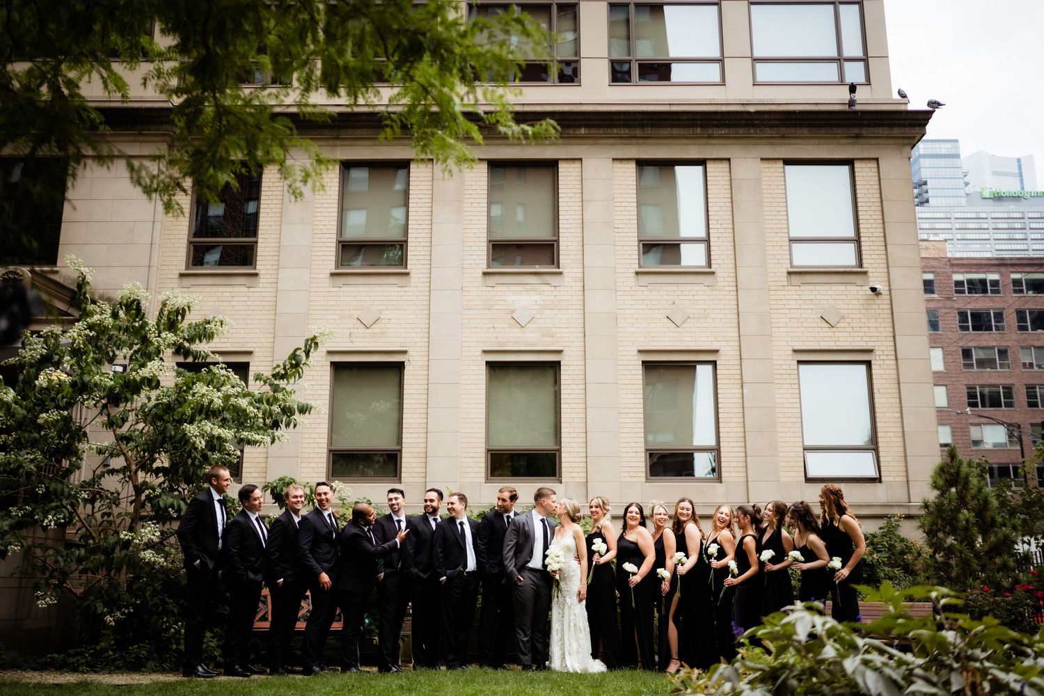 Wedding party in black and white formal attire poses for group photo outside urban building.
