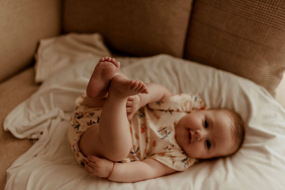 Baby in floral outfit lies on beige couch cushions showing tiny feet.