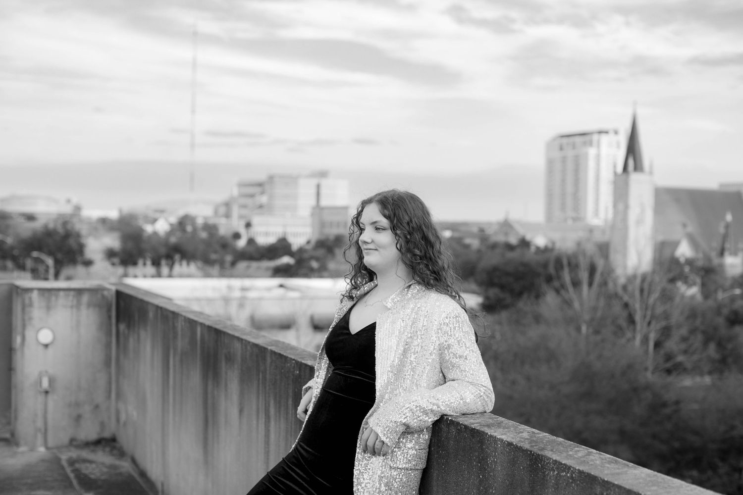 A black and white portrait of a person in a cardigan overlooking a cityscape with a church spire in the background.