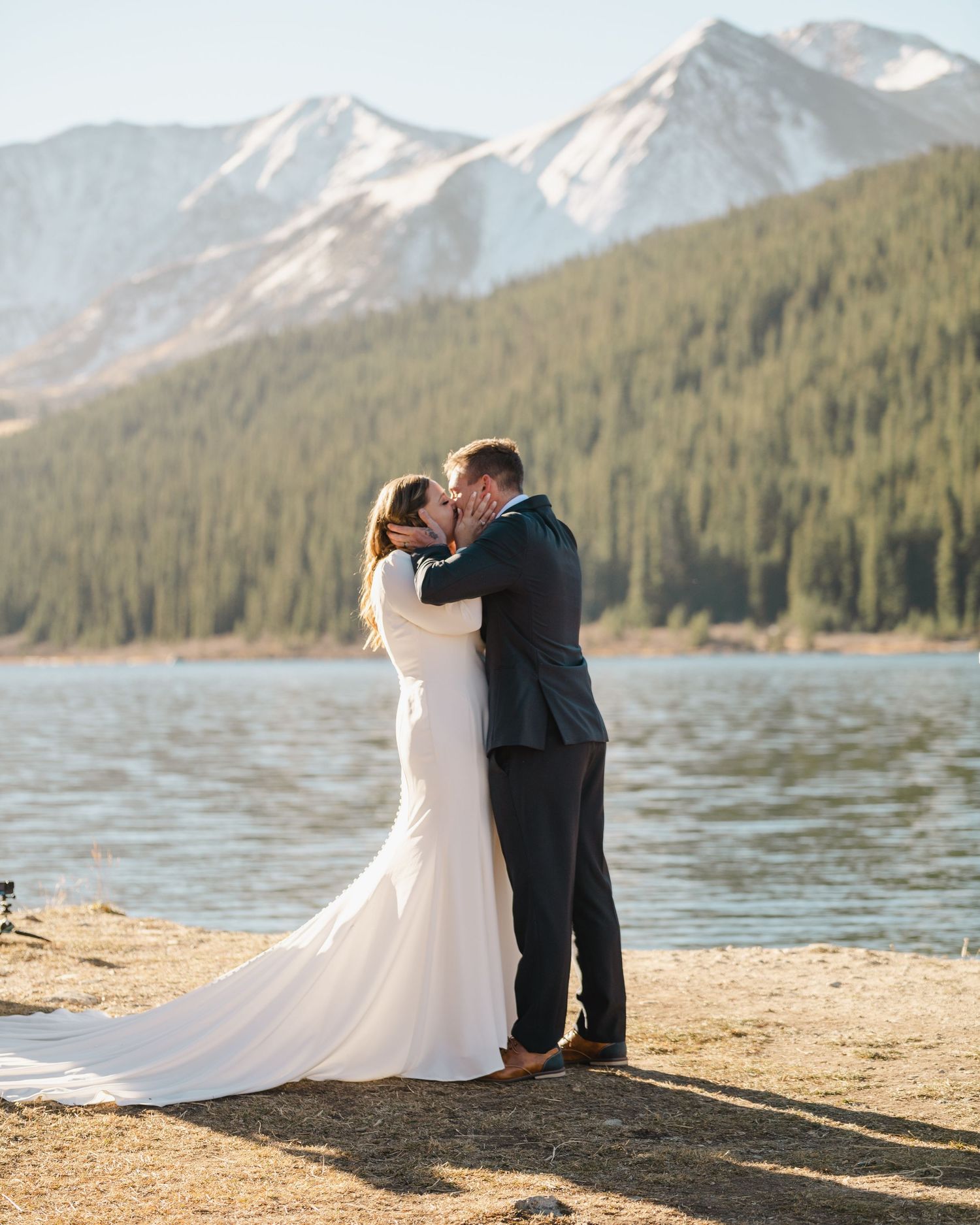 A romantic wedding kiss on a lakeshore with snow-capped mountains in the background.