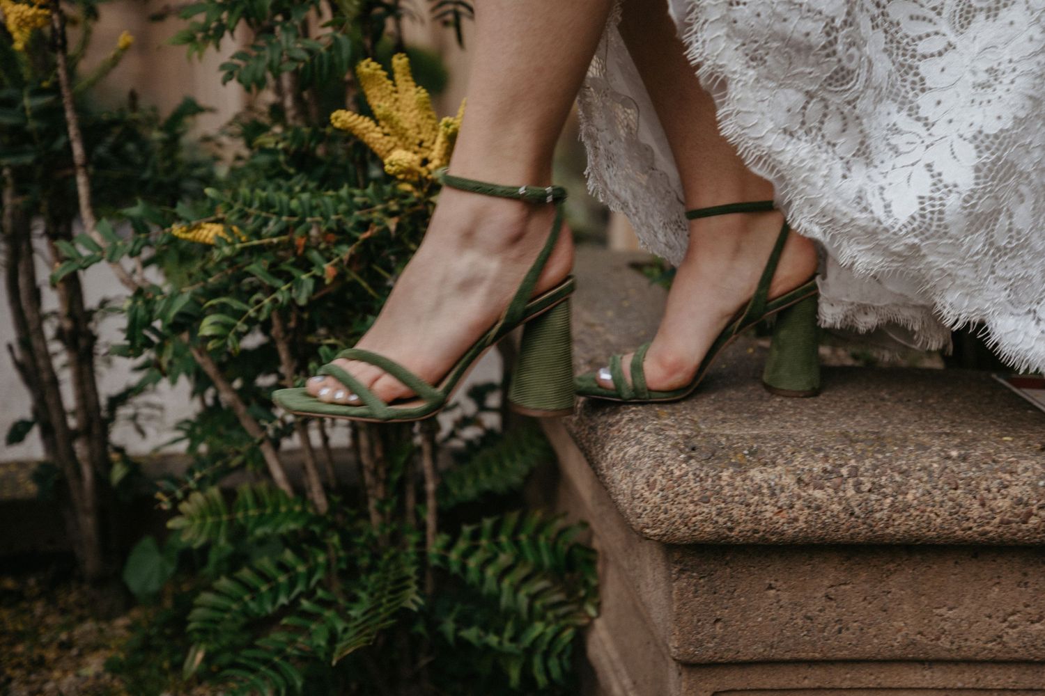 Elegant gold wedding shoes peek out from under a lace dress on stone steps with garden foliage.